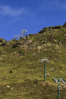 A series of chairlifts ascending a grassy, rocky hillside with a vibrant blue sky overhead. The chairlifts are unoccupied and spread in a line, supported by several tall metal towers.