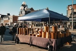 A street market scene with a canopy tent covering wooden crafts displayed on tables. The background features a row of brick buildings, indicating an urban setting. People walk by or visit the stall, creating a bustling atmosphere. A street lamp is visible, adorned with round lights.