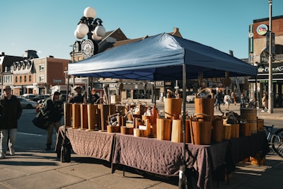 A street market scene with a canopy tent covering wooden crafts displayed on tables. The background features a row of brick buildings, indicating an urban setting. People walk by or visit the stall, creating a bustling atmosphere. A street lamp is visible, adorned with round lights.
