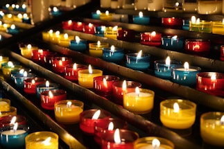 Rows of small, colorful candle holders containing lit candles are arranged neatly on metal shelves. The candles emit a warm, glowing light, creating a serene and peaceful atmosphere.