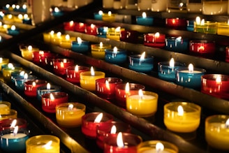 Rows of small, colorful candle holders containing lit candles are arranged neatly on metal shelves. The candles emit a warm, glowing light, creating a serene and peaceful atmosphere.