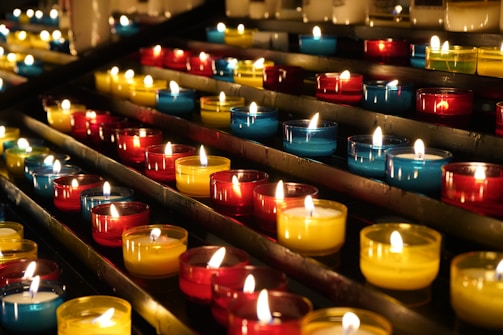 Rows of small, colorful candle holders containing lit candles are arranged neatly on metal shelves. The candles emit a warm, glowing light, creating a serene and peaceful atmosphere.