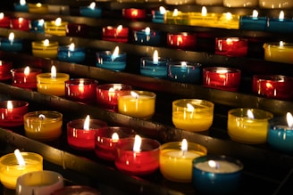 Rows of small, colorful candles are lit and arranged on black metal shelves. The candles are contained in glass holders of various colors, including red, yellow, and blue. The soft glow from the flames creates a warm and serene atmosphere.