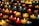 Rows of small, colorful candles are lit and arranged on black metal shelves. The candles are contained in glass holders of various colors, including red, yellow, and blue. The soft glow from the flames creates a warm and serene atmosphere.