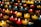 Rows of small, colorful candles are lit and arranged on black metal shelves. The candles are contained in glass holders of various colors, including red, yellow, and blue. The soft glow from the flames creates a warm and serene atmosphere.