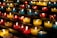 Rows of small, colorful candles are lit and arranged on black metal shelves. The candles are contained in glass holders of various colors, including red, yellow, and blue. The soft glow from the flames creates a warm and serene atmosphere.