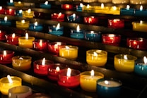 Rows of small, colorful candles are lit and arranged on black metal shelves. The candles are contained in glass holders of various colors, including red, yellow, and blue. The soft glow from the flames creates a warm and serene atmosphere.