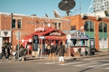 A busy urban street corner with a red BeaverTails pastry shop, signage visible, and people walking by. The brick building has large windows and satellite dishes on top. A few people are standing near the shop, possibly in line. The crosswalk and street signs are prominent, with a clear blue sky above.