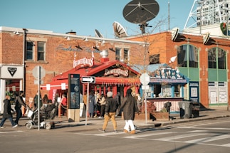 A busy urban street corner with a red BeaverTails pastry shop, signage visible, and people walking by. The brick building has large windows and satellite dishes on top. A few people are standing near the shop, possibly in line. The crosswalk and street signs are prominent, with a clear blue sky above.