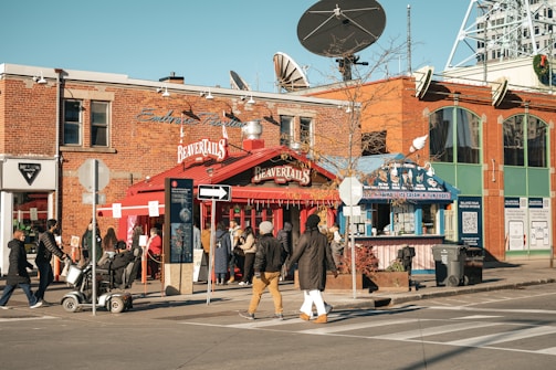 A busy urban street corner with a red BeaverTails pastry shop, signage visible, and people walking by. The brick building has large windows and satellite dishes on top. A few people are standing near the shop, possibly in line. The crosswalk and street signs are prominent, with a clear blue sky above.
