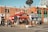 A busy urban street corner with a red BeaverTails pastry shop, signage visible, and people walking by. The brick building has large windows and satellite dishes on top. A few people are standing near the shop, possibly in line. The crosswalk and street signs are prominent, with a clear blue sky above.