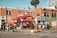 A busy urban street corner with a red BeaverTails pastry shop, signage visible, and people walking by. The brick building has large windows and satellite dishes on top. A few people are standing near the shop, possibly in line. The crosswalk and street signs are prominent, with a clear blue sky above.