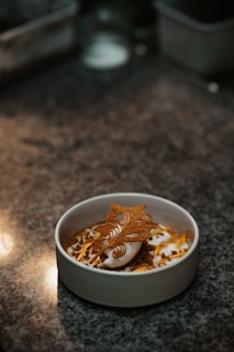 A dessert is beautifully arranged in a white bowl, placed on a granite surface. The dish includes a cream-like substance topped with an intricate caramel decoration and crumbled bits.