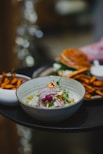 A bowl of ceviche garnished with red onion, edible flowers, and herbs is accompanied by a plate of sweet potato fries and a sandwich. The dish is artfully presented on a dark tray with a blurred background, creating an elegant dining atmosphere.