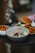 A bowl of ceviche garnished with red onion, edible flowers, and herbs is accompanied by a plate of sweet potato fries and a sandwich. The dish is artfully presented on a dark tray with a blurred background, creating an elegant dining atmosphere.