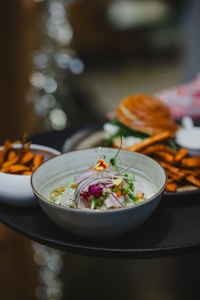 A bowl of ceviche garnished with red onion, edible flowers, and herbs is accompanied by a plate of sweet potato fries and a sandwich. The dish is artfully presented on a dark tray with a blurred background, creating an elegant dining atmosphere.