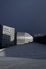 A modern urban scene with sleek architectural structures. The foreground features a large, sloped expanse of smooth, light gray paving stones. In the background, tall, contemporary buildings with a grid of windows reflect ambient light. The sky above is dark and overcast, contrasting with the illuminated buildings.