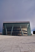 A modern architectural building with large glass facades and angled white support beams. The structure sits on a sprawling paved area with a clear geometric pattern on the ground. The design features reflective windows that mirror the surrounding environment. The sky is overcast, casting a soft light over the scene.