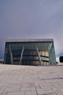 A modern architectural building with large glass facades and angled white support beams. The structure sits on a sprawling paved area with a clear geometric pattern on the ground. The design features reflective windows that mirror the surrounding environment. The sky is overcast, casting a soft light over the scene.