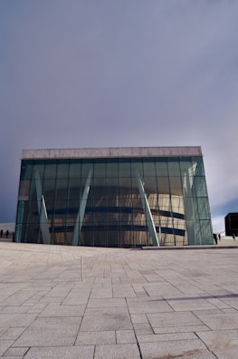 A modern architectural building with large glass facades and angled white support beams. The structure sits on a sprawling paved area with a clear geometric pattern on the ground. The design features reflective windows that mirror the surrounding environment. The sky is overcast, casting a soft light over the scene.