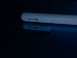 A close-up of a white stylus with the brand logo and 'pencil' printed on the side. It is lying on a reflective surface, creating a clear mirror image.