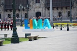 Large colorful letters forming the word LIMA are displayed prominently in an open plaza area. The background features an ornate, historic building with intricate architectural details and a row of uniformed guards standing in formation. Several people are visible, some taking photos. There are benches and streetlamps in the foreground.