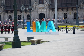 Large colorful letters forming the word LIMA are displayed prominently in an open plaza area. The background features an ornate, historic building with intricate architectural details and a row of uniformed guards standing in formation. Several people are visible, some taking photos. There are benches and streetlamps in the foreground.