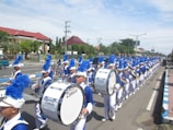 A marching band parades down a street, wearing blue and white uniforms with white hats and blue plumes. The members are playing large drums, and they form a long, straight line stretching along the road. The scene takes place in an urban area with a clear sky overhead and buildings with red roofs in the background.