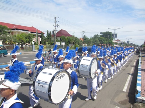 A marching band parades down a street, wearing blue and white uniforms with white hats and blue plumes. The members are playing large drums, and they form a long, straight line stretching along the road. The scene takes place in an urban area with a clear sky overhead and buildings with red roofs in the background.