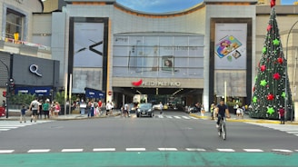 A busy urban scene featuring a large shopping mall entrance, with various advertisements for electronic products displayed on the building's facade. A tall, elaborately decorated Christmas tree stands to the right of the entrance. People are crossing the street, walking on the sidewalk, and engaging in various activities. A cyclist rides toward the viewer, while multiple vehicles are visible on the road.