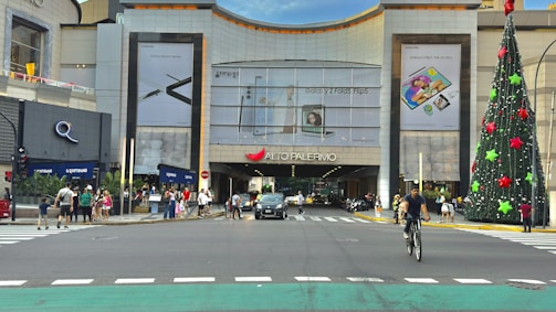A busy urban scene featuring a large shopping mall entrance, with various advertisements for electronic products displayed on the building's facade. A tall, elaborately decorated Christmas tree stands to the right of the entrance. People are crossing the street, walking on the sidewalk, and engaging in various activities. A cyclist rides toward the viewer, while multiple vehicles are visible on the road.