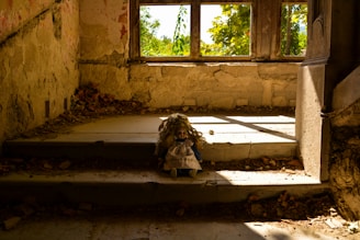 In an abandoned, dimly lit room with decaying walls and scattered debris, a doll with long curly hair and a blue dress sits alone on a stone step under a window. Natural light filters through the window, casting shadows and highlighting the doll.