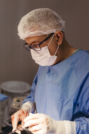 A surgeon is focused on an operation, wearing protective gear including a mask, gloves, and a hair cap. The hands are stained with blood, indicating active involvement in a surgical procedure. Medical instruments are visible, highlighting the clinical setting.