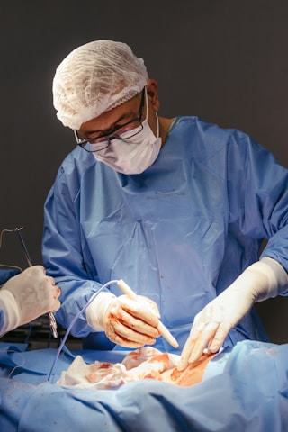 A medical professional wearing a blue surgical gown, mask, and hair net is performing surgery. The hands are gloved, and the surroundings are sterile, indicating an operating room setting. Instruments are being used, and there is a focus on a particular area of a patient's body, covered with surgical drapes.