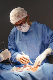 A medical professional wearing a blue surgical gown, mask, and hair net is performing surgery. The hands are gloved, and the surroundings are sterile, indicating an operating room setting. Instruments are being used, and there is a focus on a particular area of a patient's body, covered with surgical drapes.