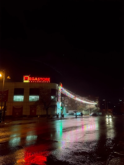 A supermarket building is illuminated at night with a bright red sign reading 'Megastore Supermarket'. Decorative lights extend from the building, reflecting colorful patterns on the wet pavement below. A few cars are visible near the entrance, and a lit streetlamp casts a warm glow over the scene.