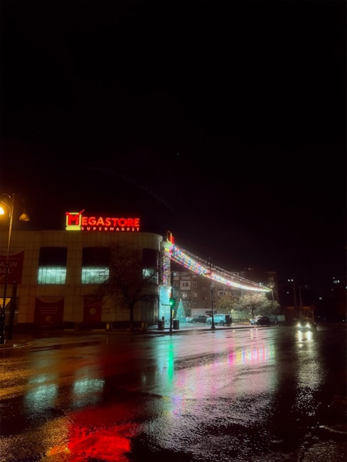 A supermarket building is illuminated at night with a bright red sign reading 'Megastore Supermarket'. Decorative lights extend from the building, reflecting colorful patterns on the wet pavement below. A few cars are visible near the entrance, and a lit streetlamp casts a warm glow over the scene.