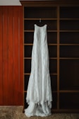 A long, white lace dress is hanging on a black hanger against a wooden shelving unit with a rich brown finish. The background includes vertical wooden panels painted orange and beige carpet at the bottom.