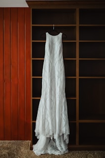 A long, white lace dress is hanging on a black hanger against a wooden shelving unit with a rich brown finish. The background includes vertical wooden panels painted orange and beige carpet at the bottom.