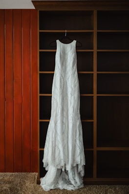 A long, white lace dress is hanging on a black hanger against a wooden shelving unit with a rich brown finish. The background includes vertical wooden panels painted orange and beige carpet at the bottom.