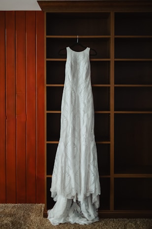 A long, white lace dress is hanging on a black hanger against a wooden shelving unit with a rich brown finish. The background includes vertical wooden panels painted orange and beige carpet at the bottom.