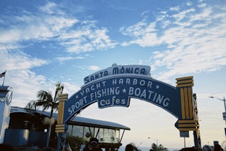 An arched sign with 'Santa Monica' written at the top stands prominently against a partly cloudy blue sky. The sign lists activities available: yacht harbor, sport fishing, boating, and cafes. Palm trees and part of a building with a glass facade appear on the left side, while several people can be seen below the sign.