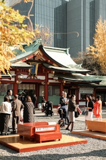 A traditional Japanese shrine with green roofs and red accents surrounded by a group of people. The setting includes a mix of modern and historical architecture, with tall contemporary buildings in the background contrasting with the traditional shrine. The people are dressed in formal attire and traditional Japanese clothing, engaging in various activities in a courtyard filled with gravel paths and wooden benches.