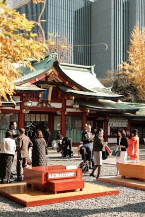 A traditional Japanese shrine with green roofs and red accents surrounded by a group of people. The setting includes a mix of modern and historical architecture, with tall contemporary buildings in the background contrasting with the traditional shrine. The people are dressed in formal attire and traditional Japanese clothing, engaging in various activities in a courtyard filled with gravel paths and wooden benches.