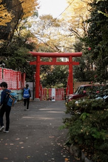 A large red torii gate stands as the focal point, surrounded by lush green foliage and trees with golden autumn leaves. Several banners or notices with red text are visible on the sides. In the foreground, two individuals with backpacks are walking on a path. A vehicle is parked to the right side, partially obscured by bushes.
