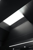 A modern and minimalistic ceiling design features a large rectangular light fixture, emitting a stark white glow. The ceiling is predominantly dark, with clean lines and subtle geometric shapes adding depth and dimension.