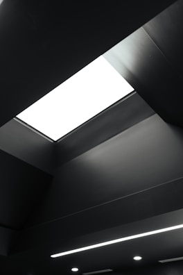 A modern and minimalistic ceiling design features a large rectangular light fixture, emitting a stark white glow. The ceiling is predominantly dark, with clean lines and subtle geometric shapes adding depth and dimension.