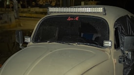 A vintage car covered in dust is parked on the side of a street at night. It has an LED light bar mounted on the roof and additional LED lights on the side mirrors. The windshield displays a sticker with the name 'Luna’s.' The car's body and windows appear worn, and there is a tarp covering part of the rear.