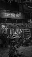 A nighttime street scene with patrons sitting outside a small bar and taco stand. The setting features string lights and a rustic wooden sign with the name 'Bar Don Pato'. Customers are seated around tables enjoying their meals, while a person stands by a grill preparing food. The background shelves hold various products, suggesting a casual dining atmosphere.