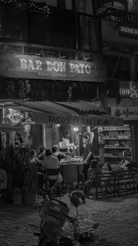A nighttime street scene with patrons sitting outside a small bar and taco stand. The setting features string lights and a rustic wooden sign with the name 'Bar Don Pato'. Customers are seated around tables enjoying their meals, while a person stands by a grill preparing food. The background shelves hold various products, suggesting a casual dining atmosphere.